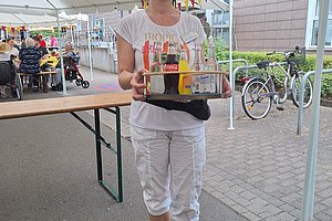 Frau mit Brille und Schirmmütze trägt Tablett mit Getränkeflaschen auf einem überdachten Außenmarkt