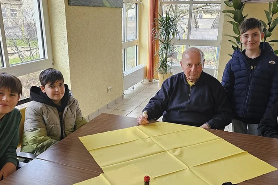 Fünf Personen sitzen und stehen um einen Tisch mit gelber Tischdecke in einem hellen Raum mit großen Fenstern und Pflanzen.