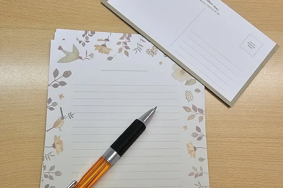 Schreibblock mit floralem Randmuster und Linien, darauf ein orange-schwarzer Kugelschreiber, daneben eine Postkarte mit Linien für Adresse und Text.