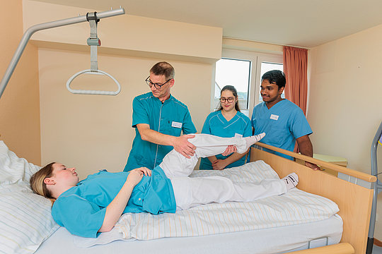 Drei in medizinischer Kleidung stehende Personen heben das Bein einer liegenden Person im Krankenhausbett