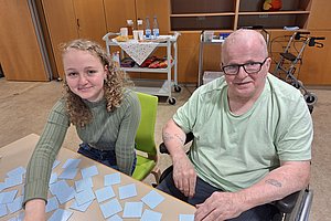 Zwei Personen sitzen an einem Tisch mit vielen Karten, eine Person im Rollstuhl, im Hintergrund Schränke und ein Rollator