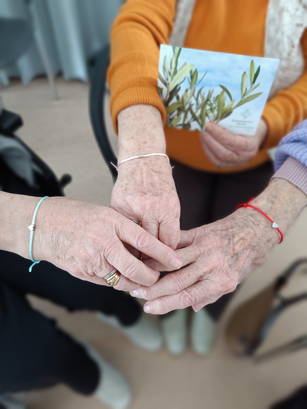 Drei ältere Hände mit dünnen Armbändern halten sich zusammen, eine Person hält eine Karte mit Olivenzweig-Motiv.