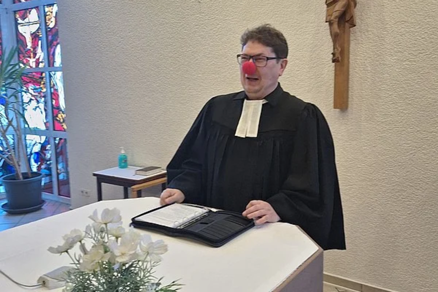 Person in schwarzer Robe mit weißem Kragen steht an einem Altar mit Blumenarrangement, im Hintergrund ein Kruzifix an der Wand.