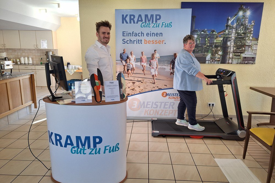 Mann steht hinter einem Messetresen mit Aufschrift 'KRAMP Gut zu Fuß', daneben Frau auf Laufband in einem Raum mit Kachelfußboden und Küchenzeile.