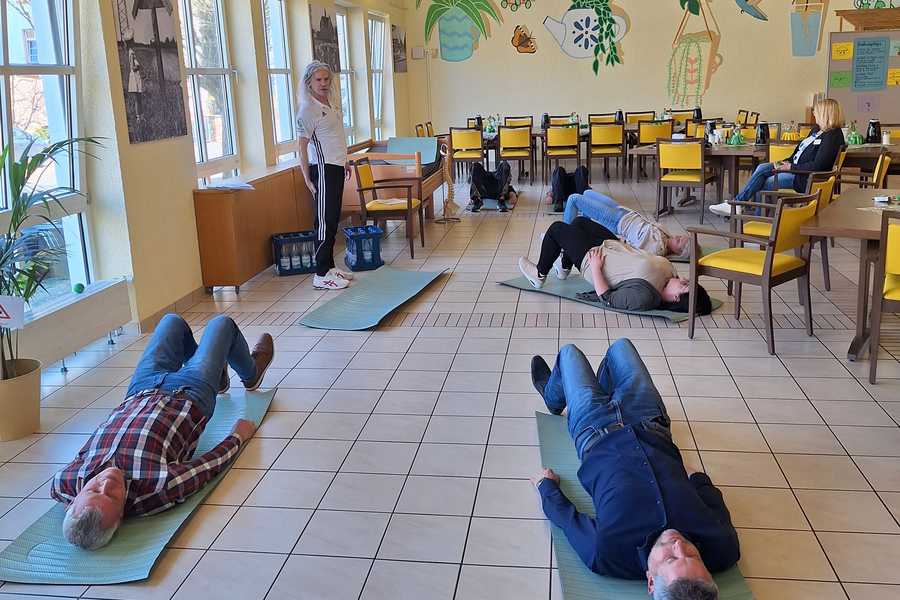 Mehrere Personen liegen auf Yogamatten in einem hellen Raum mit Pflanzenwand und Holzbalkendecke, eine Person steht an der Seite.