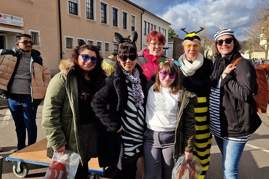 Gruppe von acht Personen, einige in Tierkostümen (Zebra, Biene), andere in Alltagskleidung, vor einem mehrstöckigen Gebäude bei sonnigem Himmel.