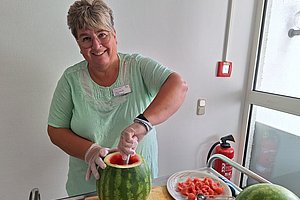 Frau mit grünem T-Shirt und Handschuhen entfernt Fruchtfleisch aus einer ausgehöhlten Wassermelone in einer Küche