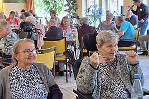 Mehrere ältere Menschen sitzen in einem hellen Raum an Tischen und unterhalten sich, eine Person hält ein Stäbchen in der Hand