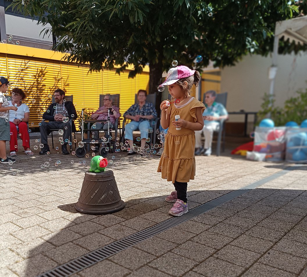 Kind in braunem Kleid und rosa Mütze steht auf gepflastertem Hof und pustet Seifenblasen, im Hintergrund mehrere sitzende Personen.