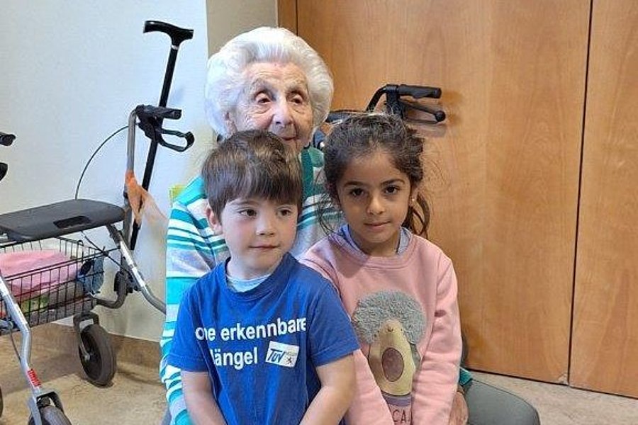 Ältere Person sitzt auf Stuhl, zwei Kinder stehen vor ihr, im Hintergrund Rollatoren und Holztüren.