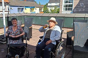 Zwei Personen im Rollstuhl sitzen auf einer Terrasse mit mehreren Stühlen und Seifenblasen in der Luft