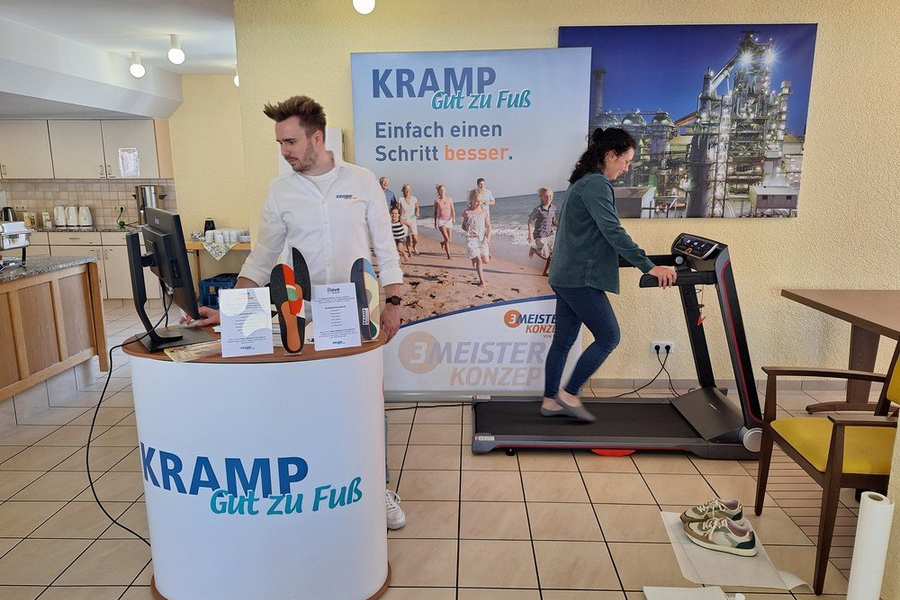 Mann steht an einem runden Messetisch mit Aufschrift 'KRAMP Gut zu Fuß', Frau geht auf einem Laufband in einem Raum mit Küchenzeile und Werbeplakat.