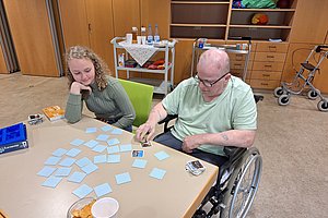 Zwei Personen sitzen an einem Tisch mit vielen Karten, eine Person im Rollstuhl, im Hintergrund Schränke und ein Rollator