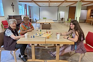 Drei Personen sitzen an einem Tisch und spielen ein Brettspiel in einem Raum mit Klavier, Kreuz an der Wand und mehreren Stühlen.