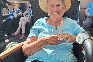 Ältere Frau mit Strohhut und gepunkteter Hose sitzt in einem Korbsessel und hält ein Glas in der Hand, im Hintergrund weitere ältere Personen auf Stühlen vor einer hellen Wand.