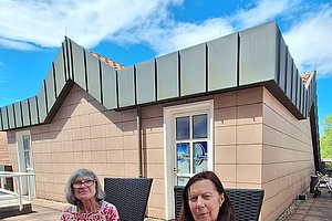 Zwei Frauen sitzen auf einem gepflasterten Hof vor einem beige verkleideten Gebäude mit grauem Dach unter blauem Himmel mit Wolken