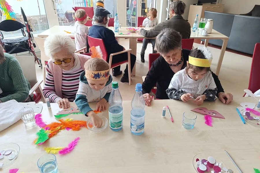 Mehrere Kinder und Erwachsene sitzen an Tischen, auf denen bunte Bastelmaterialien wie Federn, Masken und Kleber verteilt sind.