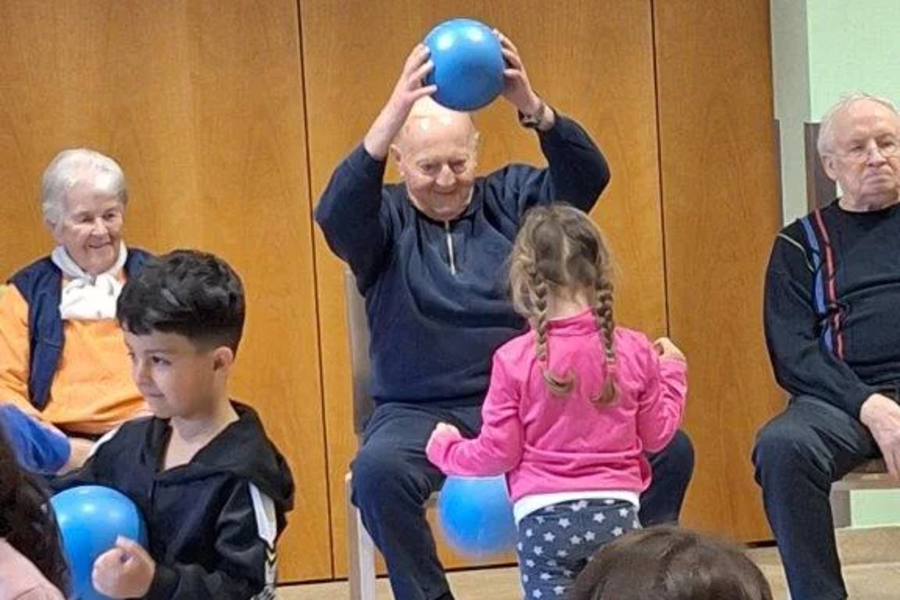 Mehrere Personen in einem Raum, zwei sitzen auf Stühlen, eine hält einen blauen Ball über dem Kopf, zwei Kinder stehen mit blauen Bällen in der Hand.