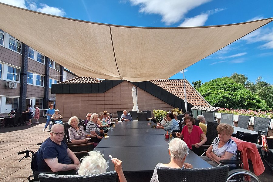 Gruppe älterer Menschen sitzt an einem langen Tisch im Freien unter einem großen Sonnensegel