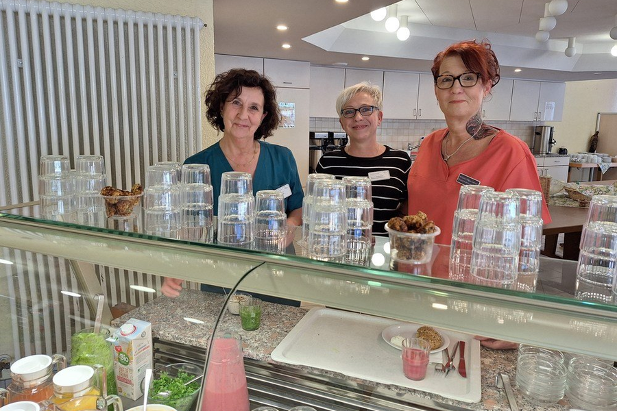 Drei Frauen stehen hinter einer Theke mit mehreren gestapelten Gläsern und kleinen Schalen mit Snacks in einem hellen Raum.