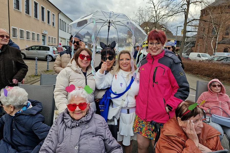 Gruppe von Menschen im Freien, darunter eine Person in weißem Kostüm mit Hasenohren und durchsichtiger Kuppelschirm über Kopf, andere tragen bunte Jacken und sitzen auf Bänken.