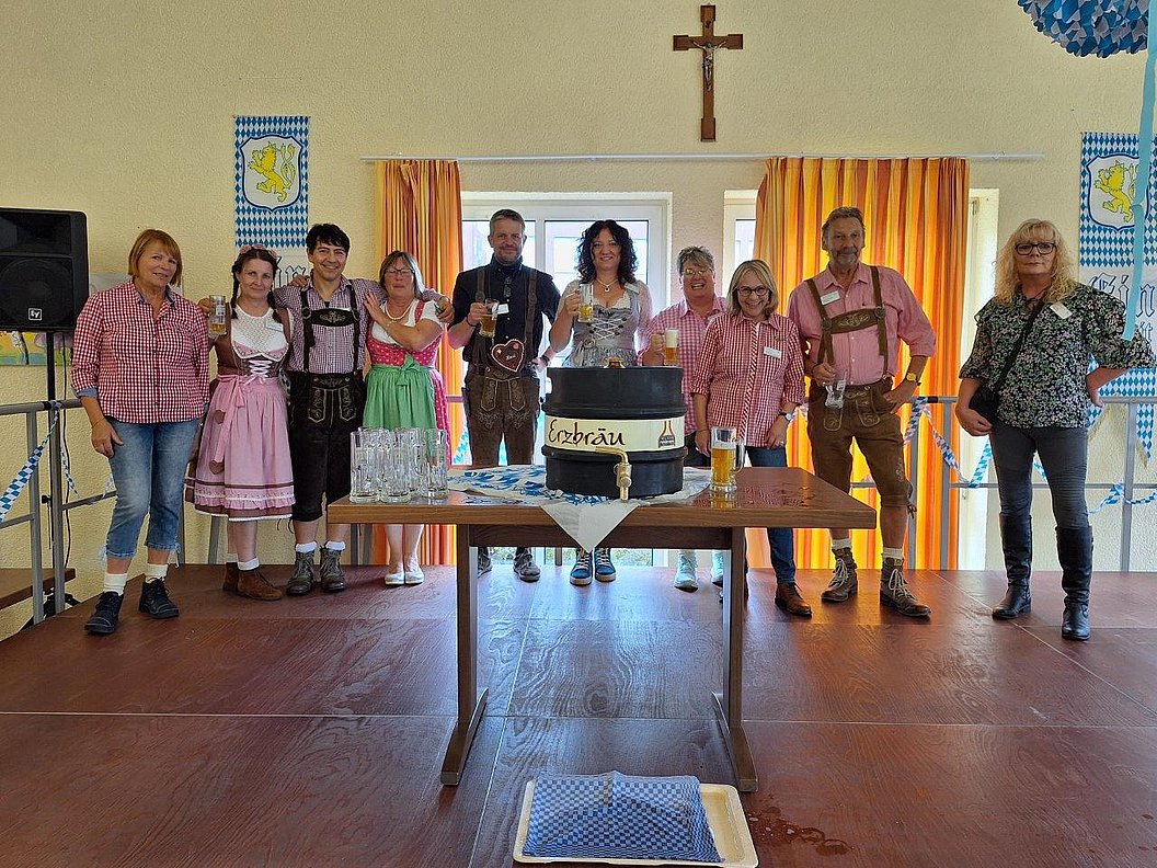Gruppe von neun Personen in Trachten vor einem Tisch mit einem Bierfass, im Hintergrund ein Kreuz und orange Vorhänge.