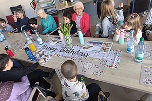 Festliche Geburtstagsfeier mit Senioren und Kindern an einem Tisch voller Getränke und Dekoration.