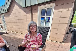 Ältere Frau mit Brille und kurz geschnittenem grauem Haar sitzt auf einer Terrasse in einem schwarzen Korbsessel und hält eine Schale mit Löffel in der Hand