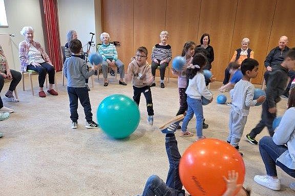 Kinder spielen mit bunten Gymnastikbällen in einem Raum, während ältere Menschen auf Stühlen an der Wand sitzen.
