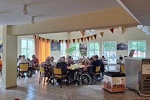 Mehrere ältere Menschen sitzen an Tischen in einem hellen Raum mit Fenstern und Girlanden, eine Person im Rollstuhl