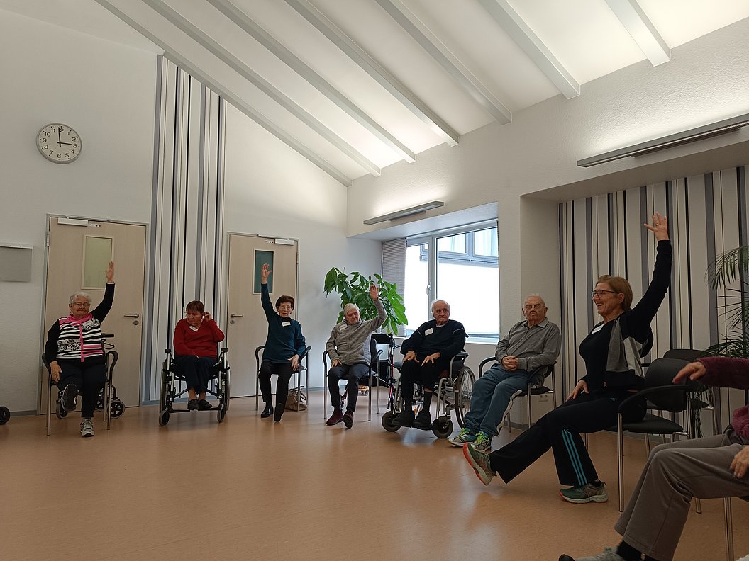 Mehrere ältere Menschen sitzen in einem hellen Raum auf Stühlen und Rollstühlen, einige heben eine Hand, eine Person streckt ein Bein nach vorne aus.