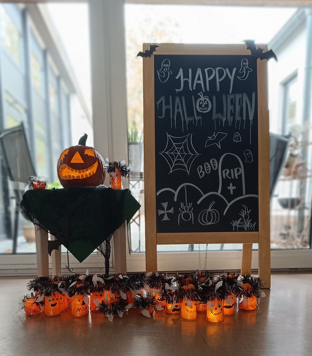 Halloween-Tafel mit handgeschriebenem Text und Zeichnungen, daneben ein Kürbis mit geschnitztem Gesicht auf einem Tisch, davor leuchtende Kürbislichter.