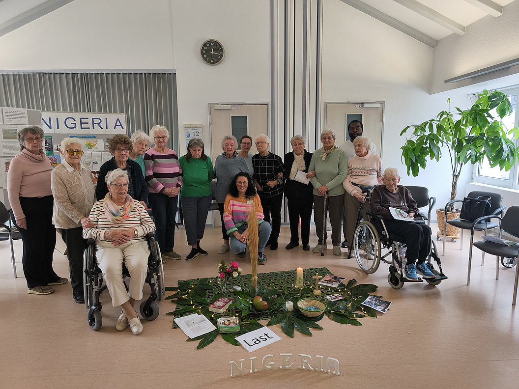 Gruppe älterer Menschen in hellem Raum um einen mit Blättern, Kerzen und Gegenständen dekorierten Bodenbereich versammelt, zwei Personen im Rollstuhl