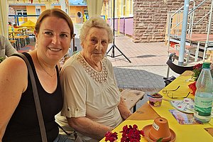 Zwei Frauen sitzen an einem gelben Tisch im Freien, auf dem eine rote Blume, ein Blumentopf und eine Wasserflasche stehen