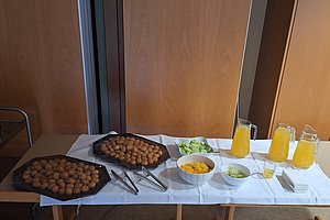 Buffet mit zwei Tabletts voller kleinen Brötchen, Schalen mit Gurkenscheiben, Käse und Salat sowie Karaffen mit Orangensaft auf einem weißen Tischtuch