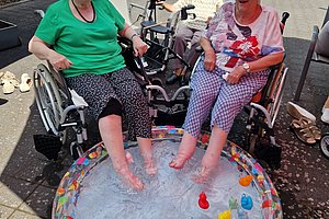 Drei ältere Frauen sitzen im Freien, zwei davon mit nackten Füßen in einem kleinen Planschbecken mit Wasser und bunten Spielzeugen