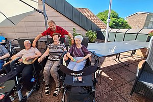 Drei ältere Menschen sitzen im Rollstuhl auf einer Terrasse, eine Person steht hinter ihnen und legt die Hände auf deren Schultern.