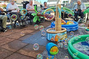 Mehrere ältere Menschen in Rollstühlen sitzen im Halbkreis im Freien vor einem Spielbereich mit aufblasbaren Pools und einer Seifenblasenmaschine, aus der viele Seifenblasen aufsteigen