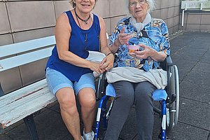 Zwei Frauen sitzen draußen auf einer Bank, eine jüngere in kurzer Hose und Top, die andere ältere im Rollstuhl mit gemustertem Hemd