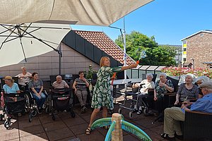 Frau in grün-weiß gemustertem Kleid steht auf Terrasse und hält einen Gegenstand vor einer Gruppe älterer Menschen, die im Schatten unter Sonnenschirmen sitzen