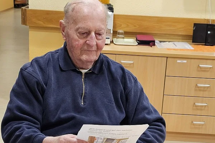 Älterer Mann sitzt an einem Tisch, hält ein Blatt Papier, auf dem Tisch liegen weitere Papiere, ein schwarzes Brillenetui und ein blauer Becher.