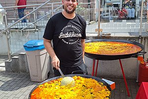 Mann mit schwarzem T-Shirt und schwarzer Kappe rührt in großer Pfanne mit gelbem und rotem Essen auf einem Gasbrenner im Freien