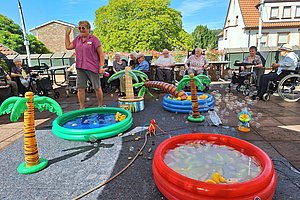 Mehrere ältere Menschen sitzen im Freien auf Stühlen und Rollstühlen um aufblasbare Planschbecken mit Palmendekor, in denen Seifenblasen schwimmen, eine Frau steht und winkt