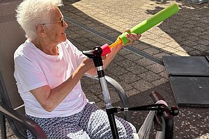 Ältere Frau mit weißem Haar sitzt im Stuhl im Freien und hält eine bunte Wasserpistole