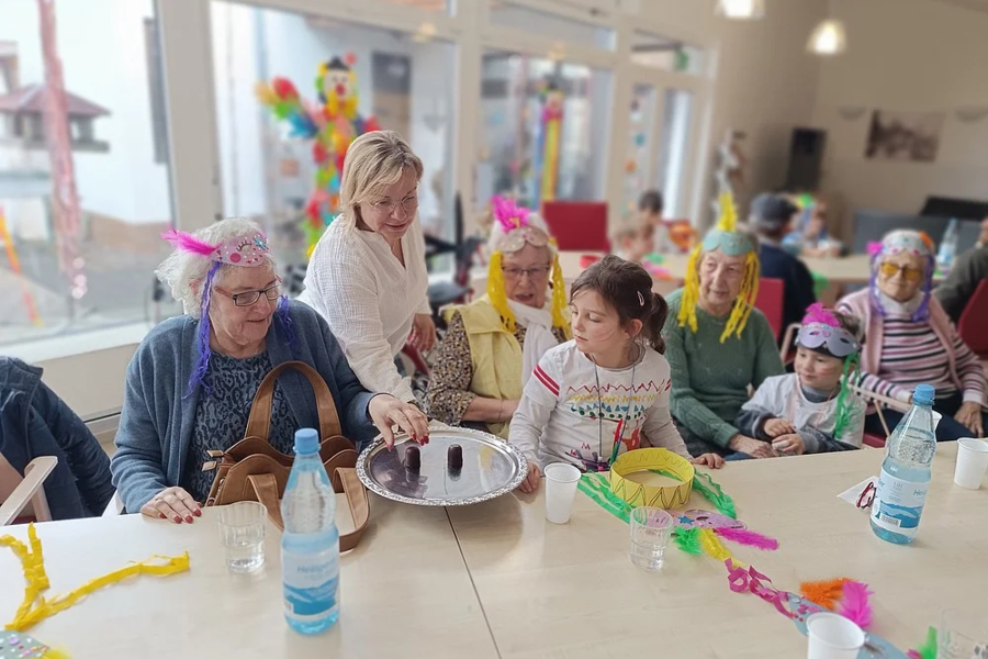Frau serviert Senioren an einem Tisch mit bunten Papiergirlanden und Wasserflaschen in einem hellen Raum.