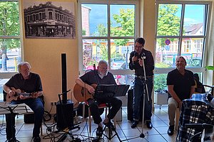 Drei Männer spielen Gitarre und ein Mann singt vor großen Fenstern in einem Raum mit Fliesenboden
