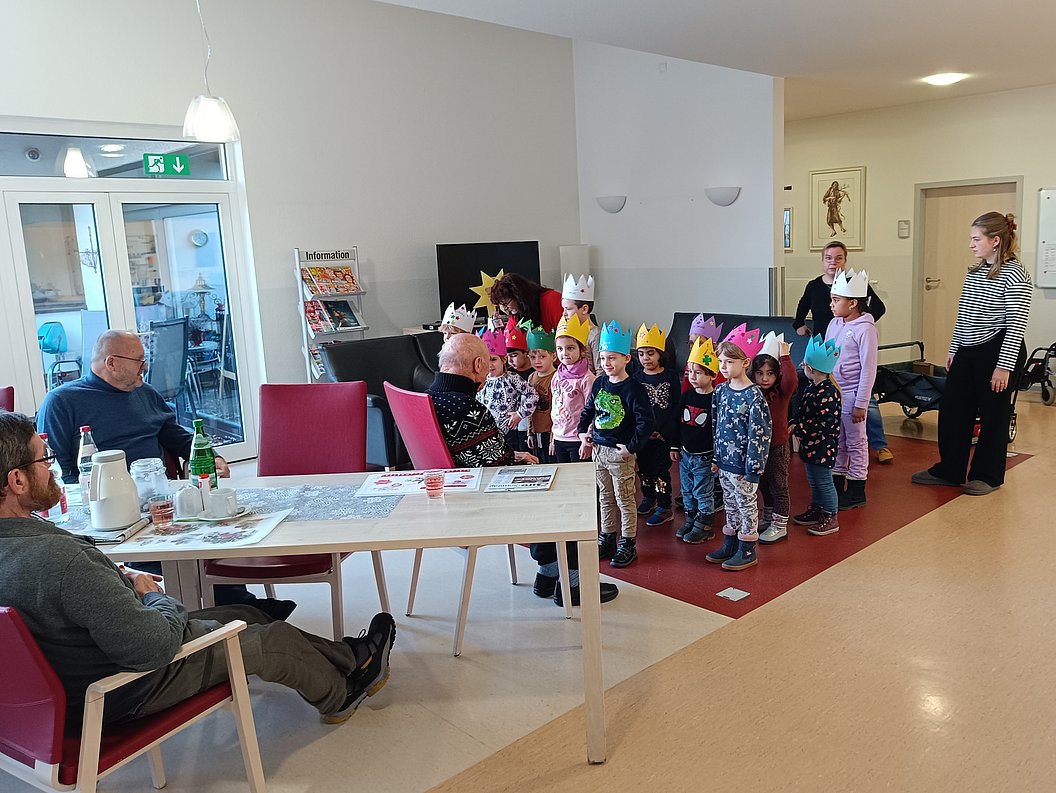 Gruppe von Kindern mit bunten Papierkronen steht in einem Raum, zwei Erwachsene sitzen an einem Tisch, eine weitere Erwachsene steht daneben.
