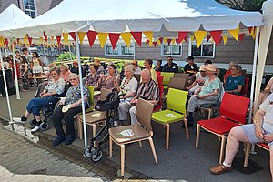 Gruppe älterer Menschen sitzt unter einem weißen Pavillon mit rot-gelben Wimpeln auf bunten Stühlen im Freien