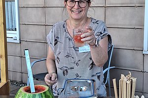Person steht hinter einem Tisch mit einem ausgehöhlten Wassermelonenbehälter, einem Metallbehälter und einem Glas mit Strohhalmen