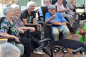 Mehrere ältere Menschen sitzen auf Stühlen und Rollstühlen im Freien auf einer Terrasse und halten Getränke in den Händen